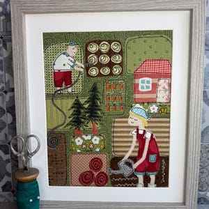 May include: A framed textile artwork showing a garden scene with two figures, a red house, and various garden plots. The frame is light grey. A pair of scissors and a spool of green thread are in the foreground, adding a craft element to the image.