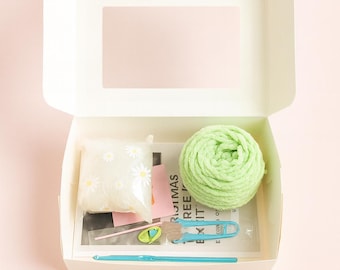 Könnte beinhalten: Eine offene weiße Box mit einem hellgrünen Garnknäuel, einem Beutel mit weißen Gänseblümchen-Verzierungen, einem blauen Häkelhaken und anderem Bastelzubehör. Die Box hat ein rechteckiges Fenster auf dem Deckel. Der Hintergrund ist ein zartes Rosa.