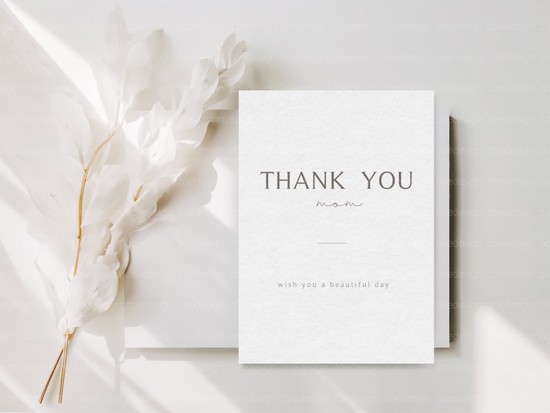 May include: A white card with the text "THANK YOU mom" and "wish you a beautiful day" printed on it. The card is sitting on a white surface with a white dried flower branch.