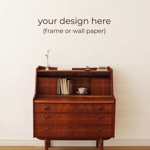 May include: A wooden desk with a drop-down front and three drawers. The desk has a light wood finish and is decorated with a vase of white flowers, a small cup and saucer, and a few books. The text "your design here (frame or wall paper)" is written on the wall behind the desk.