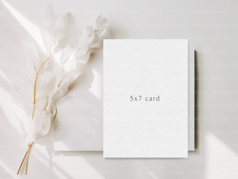 May include: A white card with the text "5x7 card" printed on it. The card is lying on a white surface with a white floral arrangement in the background.