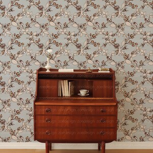 May include: A wooden desk with a drop-down front and three drawers. The desk is in front of a light blue wallpaper with a repeating pattern of brown branches and leaves.