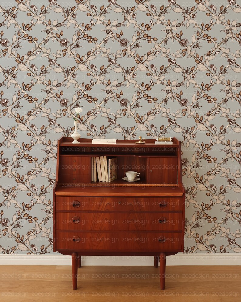 May include: A wooden desk with a drop-down front and three drawers. The desk is in front of a light blue wallpaper with a repeating pattern of brown branches and leaves.