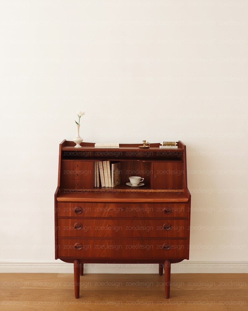 May include: A vintage wooden secretary desk with a drop-down front, three drawers, and a shelf. The desk has a light wood finish and brass hardware. It is decorated with a white flower in a vase, a small candle, and books.