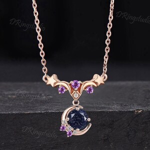 May include: A rose gold necklace featuring a crescent moon pendant set with a round, dark blue, glittery sandstone. The pendant is accented with small purple round-cut stones and clear gems, suspended from a delicate rose gold chain with star motifs.