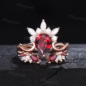 May include: A rose gold engagement ring with a pear-shaped red gemstone surrounded by smaller white gemstones. The ring has a delicate, intricate design with a band that features a vine-like pattern.
