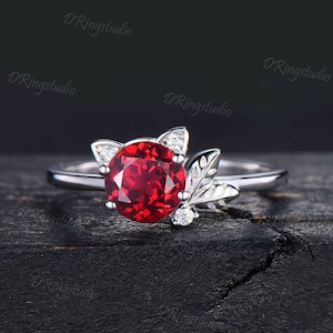 Puede incluir: Un anillo de plata con una piedra preciosa roja y un diseño de hoja. La piedra preciosa está rodeada de piedras blancas más pequeñas.