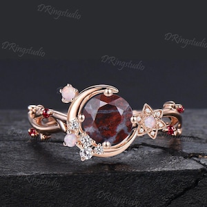 Op de afbeelding: Een roségouden ring met een grote, ronde, donkerrood gemarmerde middensteen. De band is versierd met een halve maanvorm, kleine witte opaalachtige cabochons, piepkleine heldere ronde steentjes en kleine rode steentjes op de gedraaide band.