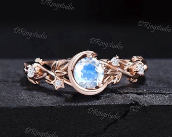May include: Rose gold ring featuring a large, round, iridescent gemstone set within a crescent moon design. The band is adorned with delicate leaves, stars, and small, sparkling accent stones. The ring has a celestial theme.