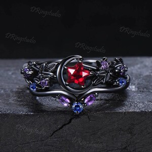 Puede incluir: Juego de anillos negros con una luna creciente que sostiene una estrella roja, con detalles florales y de estrellas. El juego incluye una banda con piedras preciosas moradas y azules. Una pieza de joyería única con temática celestial.