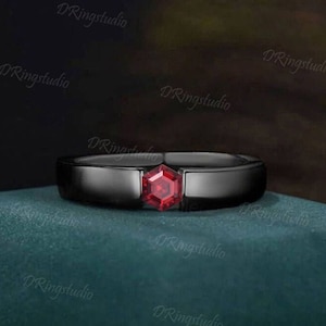 May include: A black band ring with a red gemstone in the center. The ring is on a green surface.