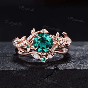 Puede incluir: Un juego de anillos de oro rosa con una gran piedra preciosa verde esmeralda redonda. El anillo está adornado con diseños florales y de hojas, y pequeñas piedras preciosas blancas. La banda también presenta pequeñas piedras preciosas blancas.