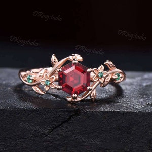 May include: A rose gold ring with a hexagonal red gemstone surrounded by delicate floral details and green gemstones.