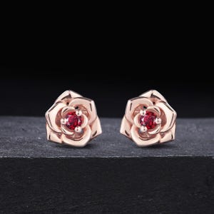 May include: A pair of rose-shaped stud earrings crafted from rose-colored metal. Each earring features a central, round-cut, deep red gemstone set within the layered, sculpted petals of the flower design, creating a floral aesthetic.