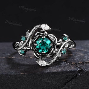 May include: A black ring featuring a green gemstone set in a rose-shaped design. The ring has a snake-like band with a smaller white gemstone. The ring is surrounded by smaller green gemstones.