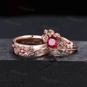 Op de afbeelding: Een paar trouwringen van roségoud met een bloemmotief. De ring van de vrouw heeft een grote rode edelsteen omringd door kleinere witte edelstenen. De ring van de man heeft een kleinere rode edelsteen.