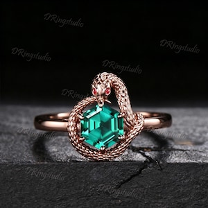 May include: A rose gold ring featuring a hexagonal green gemstone held by a coiled snake with textured scales and small red eyes. The snake's head is positioned at the top of the stone, and the band is smooth. The text DRingstudio is visible.