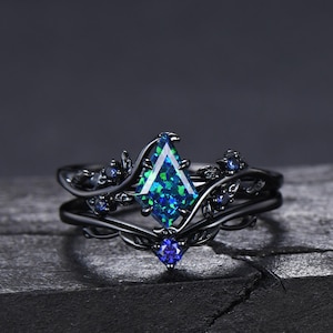 May include: Black ring with a large, green and blue iridescent gemstone set in a delicate, black metal vine design. The ring is accented with smaller blue gemstones.