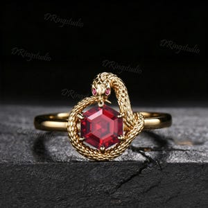 May include: A gold ring featuring a textured snake coiled around a central hexagonal red gemstone. The snake has small red eyes and its body forms a circular frame around the faceted stone, which is set on a smooth, polished gold band.