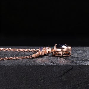 May include: A rose gold-toned necklace featuring a dark blue, glittery, round-cut stone pendant set in a metal frame. The chain is composed of delicate, interlocking oval links, and the pendant is accented with small, light-colored, clear stones.