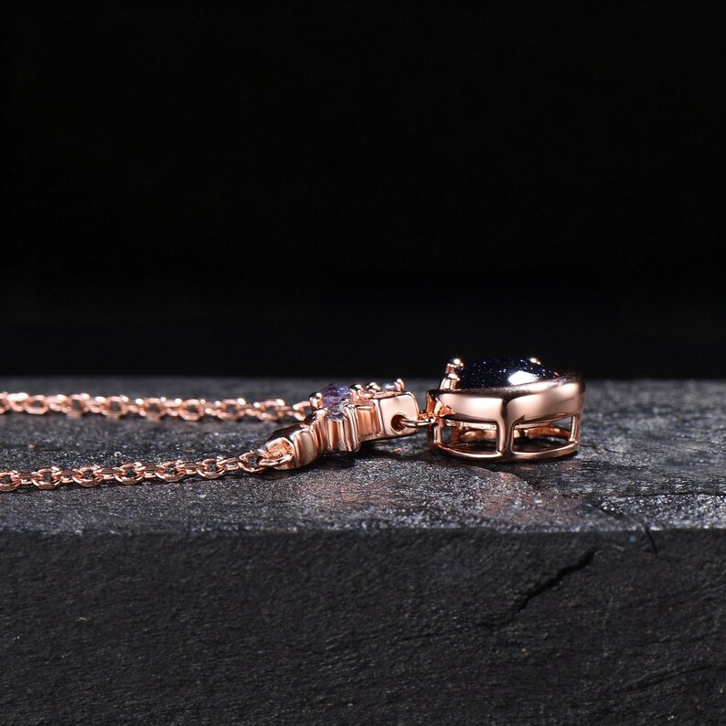 May include: A rose gold-toned necklace featuring a dark blue, glittery, round-cut stone pendant set in a metal frame. The chain is composed of delicate, interlocking oval links, and the pendant is accented with small, light-colored, clear stones.