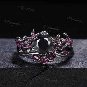 May include: A black gemstone ring with a floral design. The ring features a large, round black stone at the center, surrounded by smaller pink gemstones and floral accents. The band is dark-colored.