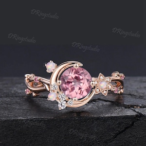 May include: A rose gold ring featuring a large round pink center stone set within a crescent moon shape. The band is adorned with small white opals, clear round accent stones, and tiny pink stones arranged in a delicate, vine-like floral pattern.