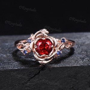 May include: A rose gold ring with a red gemstone set in a rose design. The ring is accented with smaller blue gemstones.