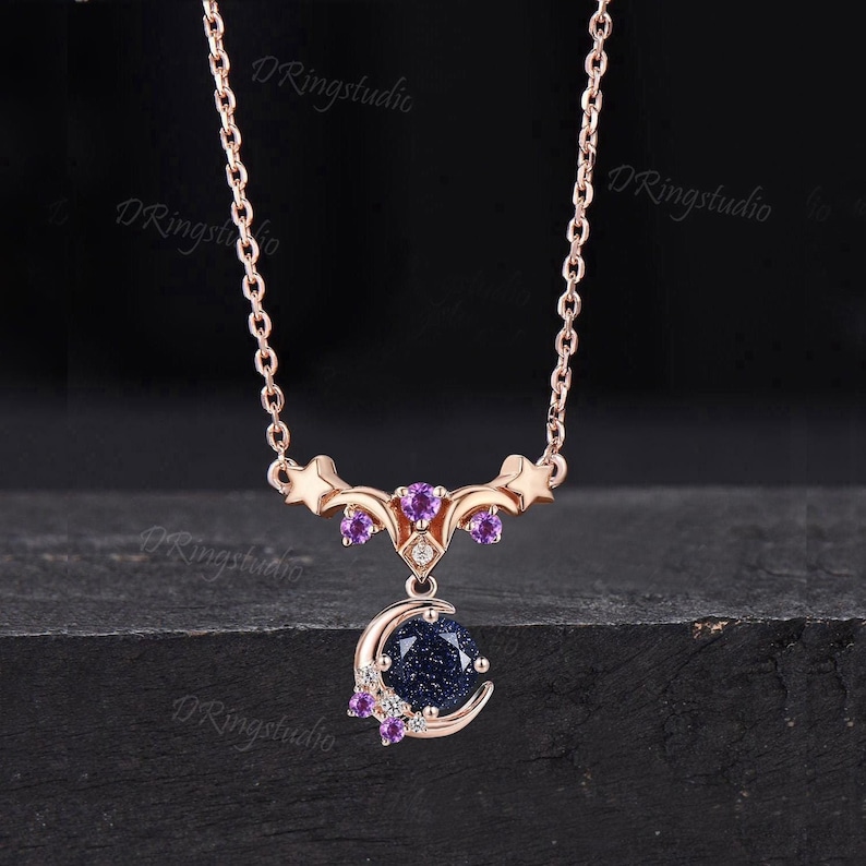 May include: A rose gold necklace featuring a crescent moon pendant set with a round, dark blue, glittery sandstone. The pendant is accented with small purple round-cut stones and clear gems, suspended from a delicate rose gold chain with star motifs.