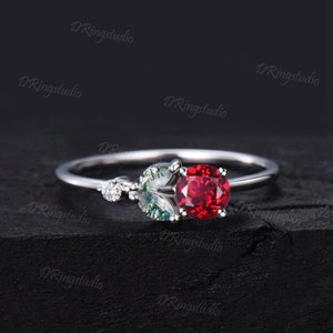 May include: A silver ring with a red gemstone and a green and white gemstone. The ring is set on a dark background.