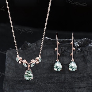 May include: A rose gold necklace and matching earrings set. The necklace features a teardrop-shaped pendant and leaf accents with green and white stone inlays. The earrings have a similar design, with teardrop-shaped stones.