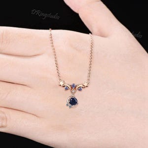 May include: A rose gold necklace featuring a celestial-themed pendant with a round dark blue glittery stone, a crescent moon accent, and small star motifs. The pendant is adorned with tiny purple and clear stones, suspended from a delicate chain.