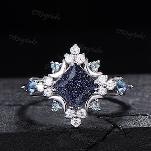 May include: A silver ring with a square, dark blue center stone with a sparkling effect. The ring features smaller white and blue accent stones, arranged in a decorative pattern around the center stone. The ring is set against a dark background.