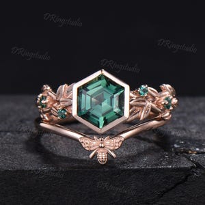 May include: A rose gold ring set featuring a central hexagonal green gemstone. The band is adorned with leaf motifs and small green accent stones. A separate V-shaped band features a small bee detail at the center, completing the nature-inspired design.