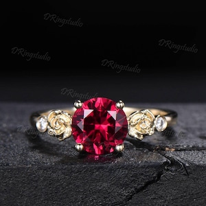 Könnte beinhalten: Ein goldener Ring mit einem großen, runden, facettierten Rubin. Der Ring hat rosenförmige Verzierungen und kleine, klare Edelsteine. Der Ring ist vor einem dunklen Hintergrund platziert.