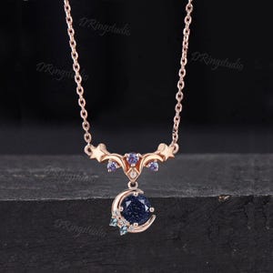 May include: A rose gold necklace featuring a celestial-themed pendant with a round, dark blue glittery stone set within a crescent moon. The design includes small stars, light purple accent stones, and clear crystals on a delicate chain.