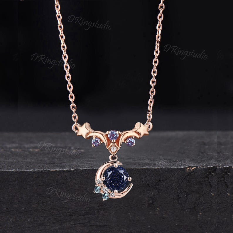 May include: A rose gold necklace featuring a celestial-themed pendant with a round, dark blue glittery stone set within a crescent moon. The design includes small stars, light purple accent stones, and clear crystals on a delicate chain.