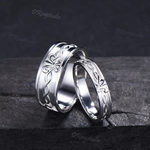 Könnte beinhalten: Zwei silberfarbene Ringe mit polierter Oberfläche und einem durchgehenden geprägten Muster aus verschlungenen Linien und stilisierten Blumenmotiven. Der breitere Ring steht hinter einem schmaleren, passenden Ring, beide mit reflektierender Oberfläche.