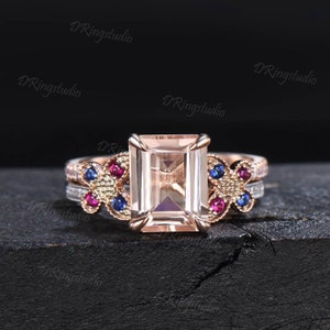 May include: A rose gold engagement ring with a large, rectangular, light pink gemstone. The ring has two bands, one with a delicate diamond pattern and the other with a butterfly design featuring red, blue, and white gemstones.