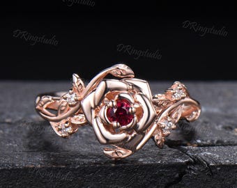 May include: A rose gold ring featuring a central deep red gemstone shaped like a flower bloom, surrounded by metallic rose-shaped petals and small clear accent stones along the band. The text DRingstudio is visible in the background.