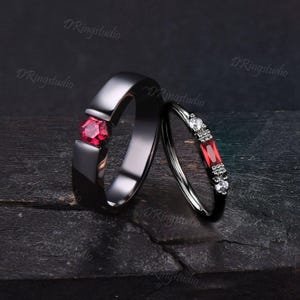 May include: Two black rings with red gemstones. The larger ring has a single, square-shaped red gemstone. The smaller ring has three rectangular red gemstones with smaller white gemstones on either side.
