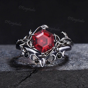 May include: A silver ring featuring a red gemstone set in a hexagonal shape. The ring has a delicate, intricate design with black accents.