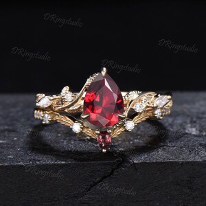 May include: A pear-shaped red gemstone ring set in gold with a delicate floral design and smaller white gemstones. The ring is part of a matching set with a smaller band.