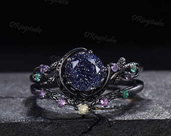 May include: A black metal ring set featuring a round, dark blue, glittery sandstone center stone. The band is shaped like a crescent moon and vine, accented with small, round, multi-colored gemstones in shades of purple, green, and yellow.