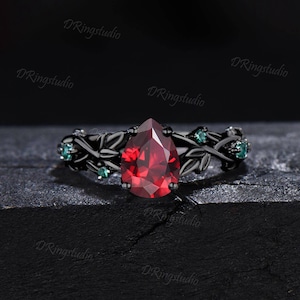 May include: Black engagement ring with a pear-shaped red gemstone and green accents. The ring features a delicate vine design.
