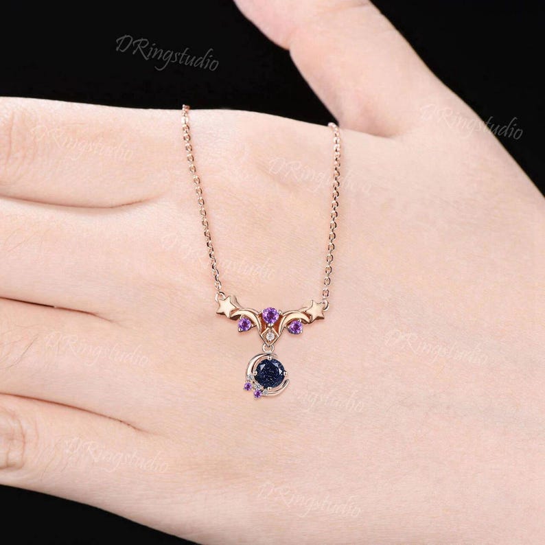 May include: A rose gold necklace featuring a central pendant with a round dark blue glittery stone, a crescent moon shape, and small purple accent stones. The metal frame includes star motifs and connects to a delicate rose gold chain.