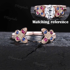 May include: A rose gold ring with two butterfly designs. Each butterfly has red and blue gemstones and a pave setting. The butterflies are facing each other and are connected by a band. The ring is on a dark wooden surface. Matching reference.