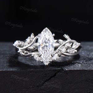 Op de afbeelding: Een zilverkleurige ring met een grote marquise-geslepen middensteen en kleinere ronde en marquise-geslepen accentstenen in een rankachtig patroon langs de band. Het metaal heeft een gepolijste afwerking en het ontwerp bevat bladmotieven.