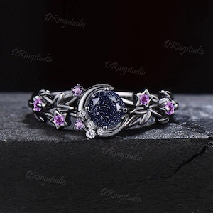 Könnte beinhalten: Ein silberner Ring mit einem dunkelblauen Edelstein in der Mitte, verziert mit einem Mond und Sternen. Der Ring ist mit kleinen lila Edelsteinen und weißen Akzenten verziert. Der Ring hat ein himmlisches Thema.