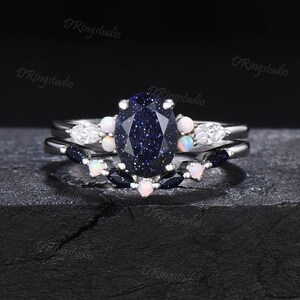 May include: A three-piece ring set featuring a large oval-shaped blue gemstone with a sparkly finish. The center stone is surrounded by smaller white gemstones and blue gemstones. The ring is made of silver.